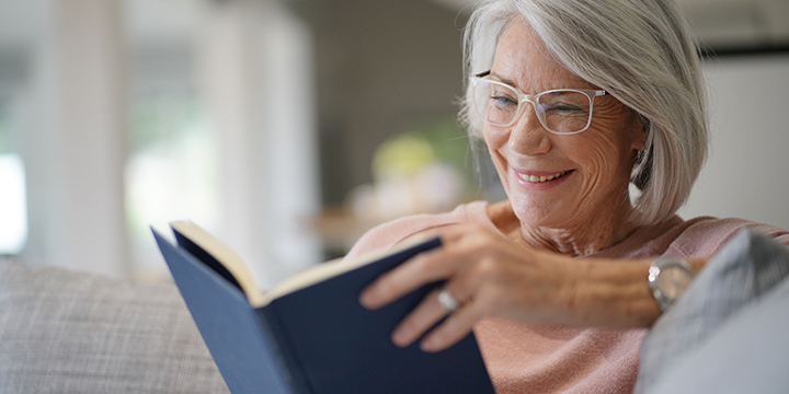 An older woman sitting on a couch, reading a book and smiling.