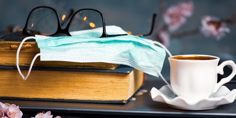 Photograph of a pair of glasses sitting on a book with a facemask next to a cup of tea