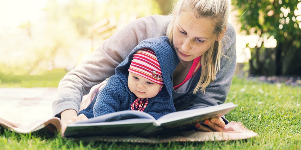A photograph of a parent and child reading a book on a blanket outside.