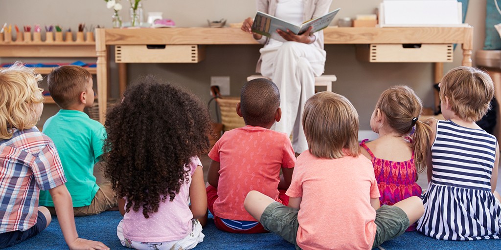 A group of preschool aged children sitting on the floor photograph from behind. An adult is reading a book in front of them.