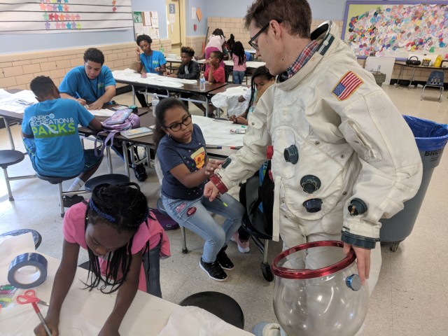 Many students are working on their space suit designs, in the center ground a young student examines the suit Spaceman Rick is wearing for design inspiration.
