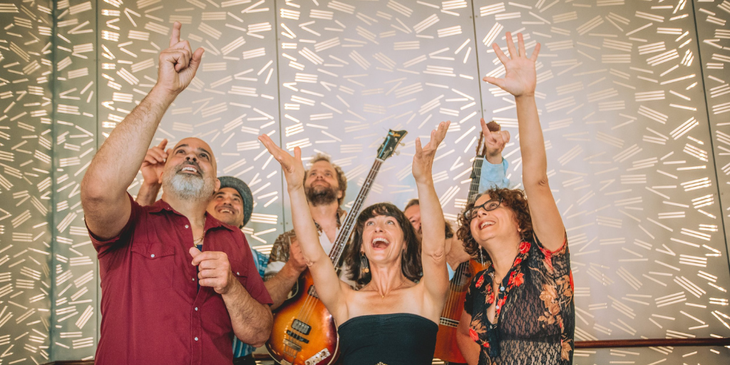 Six band members stand together in a group. Two of them carry guitars. They all stare upward, smiling and raising their arms. 