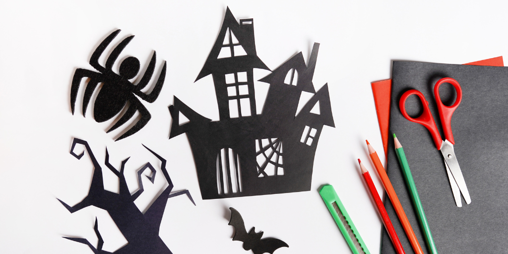 A close-up photograph of construction paper, scissors, and colored pencils sitting on a white table. Next to them are black paper cut outs of a bat, spider, spooky tree, and haunted house. 