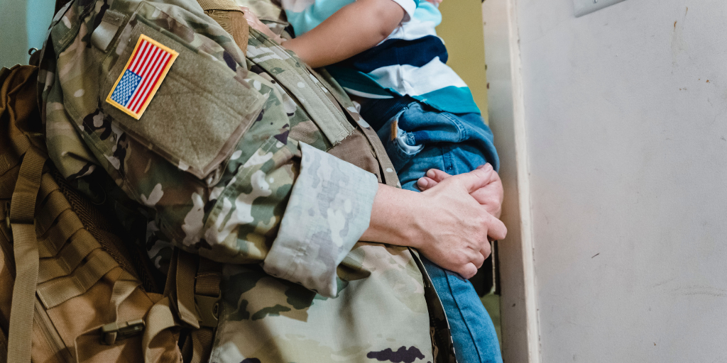 A close up image shows the torso of an adult wearing a camouflage jacket holding a toddler in a striped shirt and jeans.