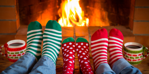 Three pairs of feet in colorful striped and polka-dotted socks sit perched in front of a fireplace. The feet are large and small. Two coffee mugs sit on the fireplace. 