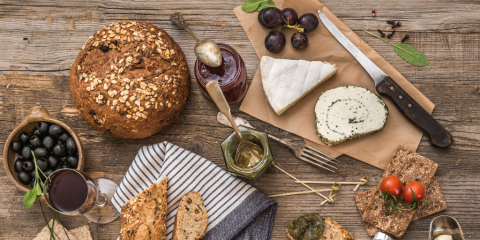 A photograph taken from a bird's-eye-view features a rustic wooden table topped with a spread of bread loaves, different cheeses, olives, tomatoes, spreads, and napkins.