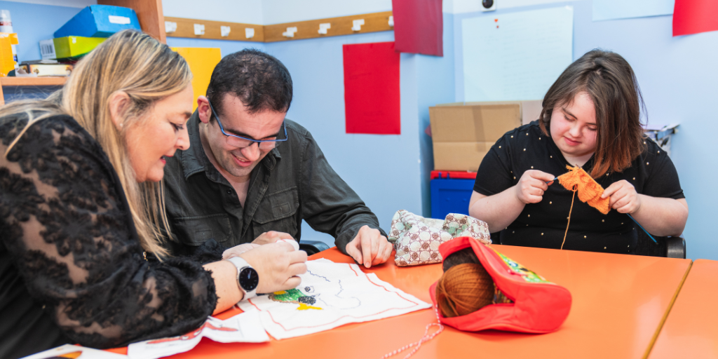 Three adults sit side-by-side at a table while knitting and coloring. One of the adults assists the other as he colors.  