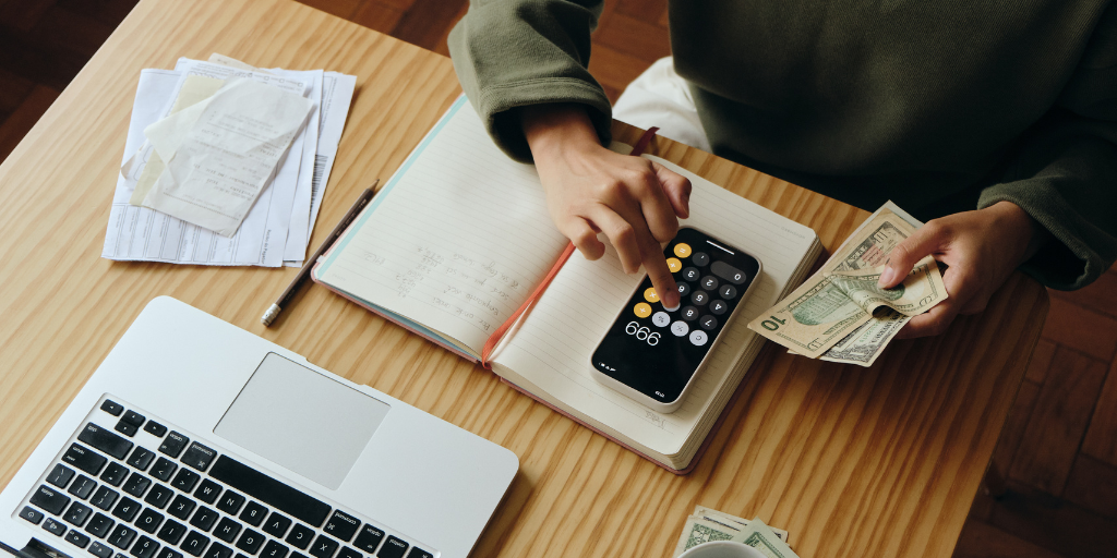 A bird's-eye-view shows a young person holding cash in one hand while using their phone calculator with the other. Beneath their phone is a notebook and pencil. A laptop and other documents are visible on the table. 