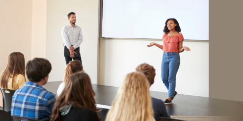 A young woman gives a presentation in front of a group of teens. The young woman stands in front of a projector screen, and a young man stands off to the side listening.  