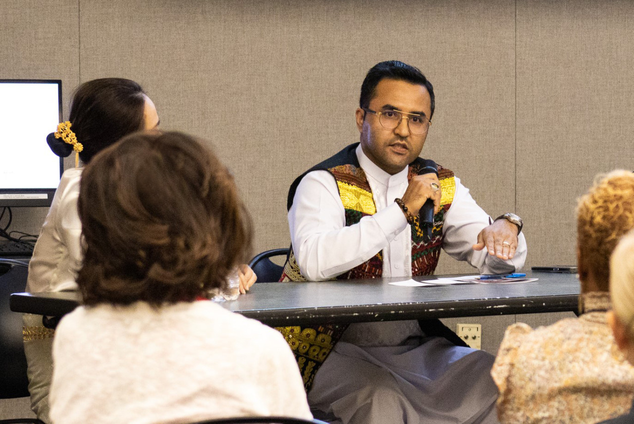 A man of Afghan descent sits at a table in front of a room full of people.. He shares his story about immigrating to the United States from Afghanistan.