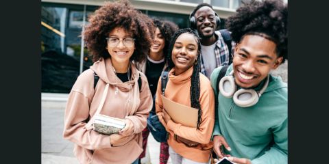 Five teen students stand in a group and smile toward the camera. They carry notebooks, backpacks, and some wear headphones. 