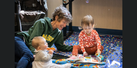 A smiling older woman lays on her side on the library floor. To her right sits her grandchild, a baby, playing with a rattle. To her left sits her other grandchild, a toddler, who looks at a book on the floor in front of her.