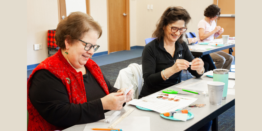 Two women wearing glasses sit side-by-side at a table, smiling, as they both craft holiday ornaments.
