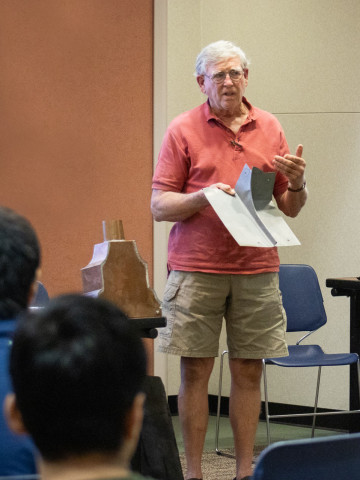 A presenter stands at the front of a meeting room. He showcases traditional roofing materials. In the foreground we see the backs of teens' heads as they sit in the audience listening.