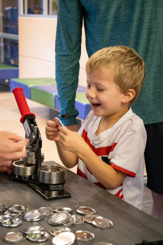 A young boy smiles as he holds the button he just finished making with the button maker in front of him.