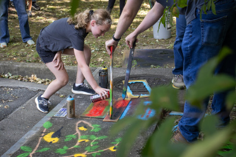 A teen bends over to spray paint a stencil on the concrete of a storm drain. The drain features artwork of birds and plants.