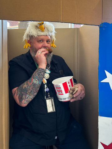 A man wearing a chicken hat eats fried chicken from a bucket. 