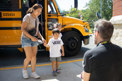 A woman and young child walk off of a school bus and are greeted by a library staff member who kneels on the ground with a prize. 