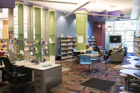 A wide-angle photograph of the Teen Room at a library. In the foreground is a reference desk, beyond that are tables and chairs where teens can work and relax, and in the background is a video gaming area and tv console. Along the walls of the room are book shelves. 