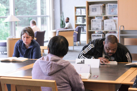 Three adults with special needs sit together at a table in the library and read.