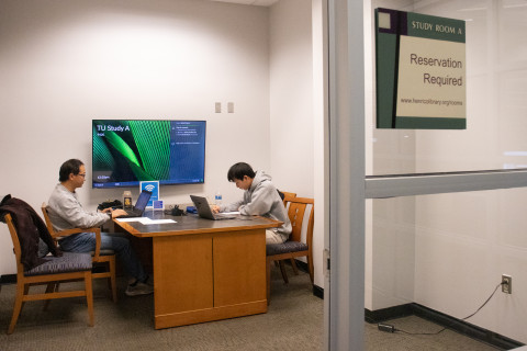 An adult and teen sit at a table in a library study room working on their laptops.