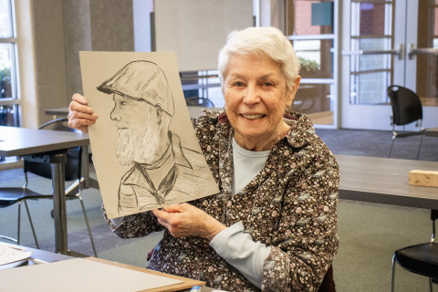 An older adult smiles at the camera while holding up her charcoal portrait of an older gentleman.