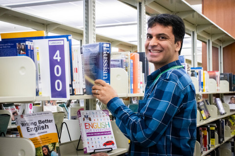 A man wearing a blue plaid shirt places a large Russian dictionary on a library shelf. He smiles at the camera.
