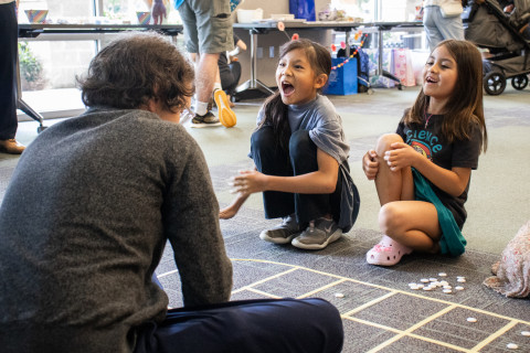An adult and two children sit on the floor and play a Vietnamese game. Masking tape marks a game board on the floor. The two children are smiling and laughing.