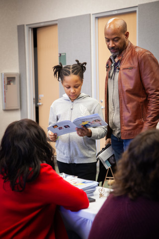 An adult and tween stand and flip through a book together. In front of them is the author sitting at a table with her books and other promotional materials. 