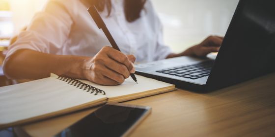 Person working on a laptop and writing on a notepad